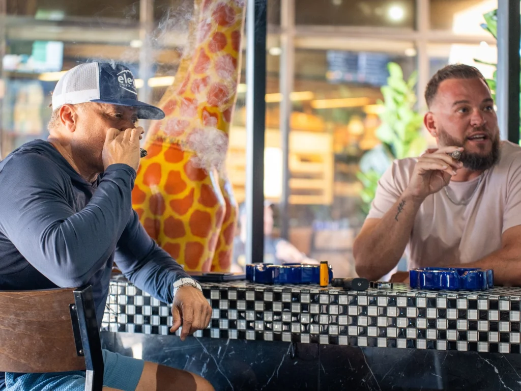 Two men smoke cigars at the checkered-tile bar at Fox Cigar Bar Gilbert, a bright giraffe statue behind them