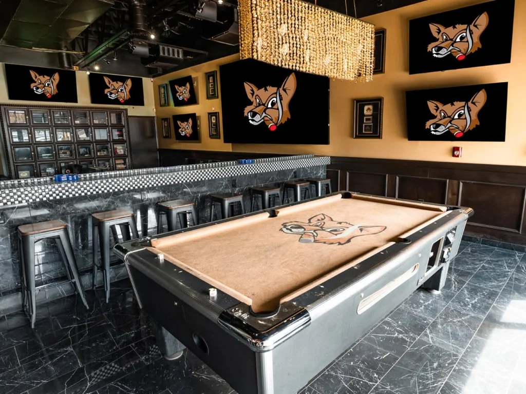 Pool table with a fox logo on the felt at Fox Cigar Bar Gilbert, bar stools, a crystal chandelier and fox-logo screens behind
