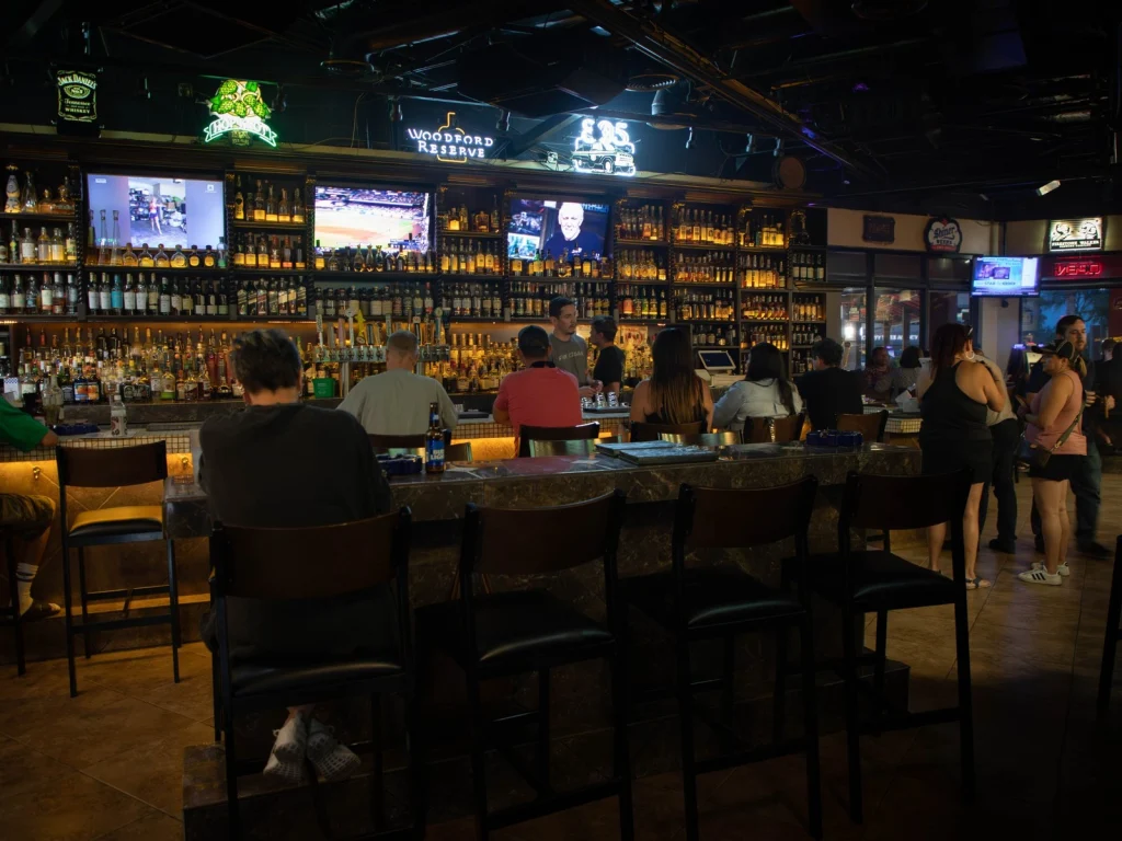 Guests seated and standing at the main bar of Fox Cigar Bar Gilbert in the evening, neon signs and a lit back bar of bottles