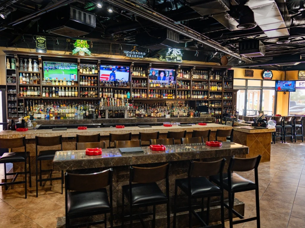 Wide view of the main bar at Fox Cigar Bar Gilbert, with a tiled counter, leather stools and a back bar lined with bottles and TVs