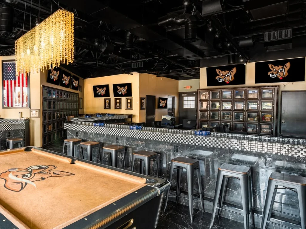 Pool table with a fox logo and metal stools along a checkered bar at Fox Cigar Bar Gilbert, cigar lockers and a chandelier behind