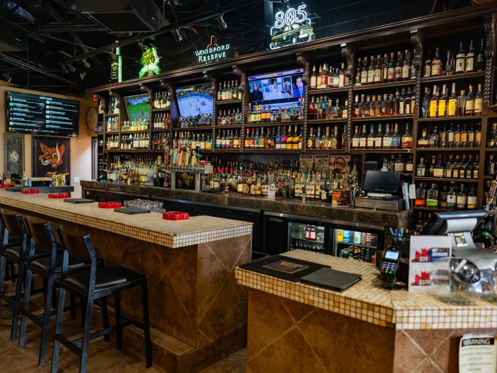 Back bar wall at Fox Cigar Bar Gilbert packed with whiskey bottles, neon signs, TVs and a draft beer menu above a tiled counter