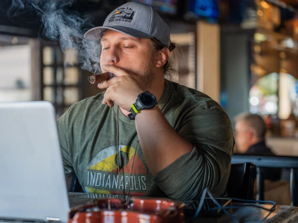 A guest in a cap enjoys a cigar with a laptop open and a clay ashtray at Fox Cigar Bar Scottsdale