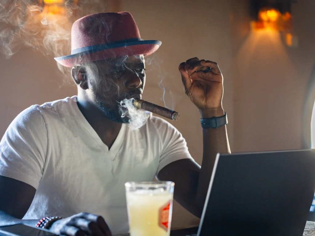 A man in a red fedora smokes a cigar beside a laptop and a cold drink in the warm light of Fox Cigar Bar Scottsdale