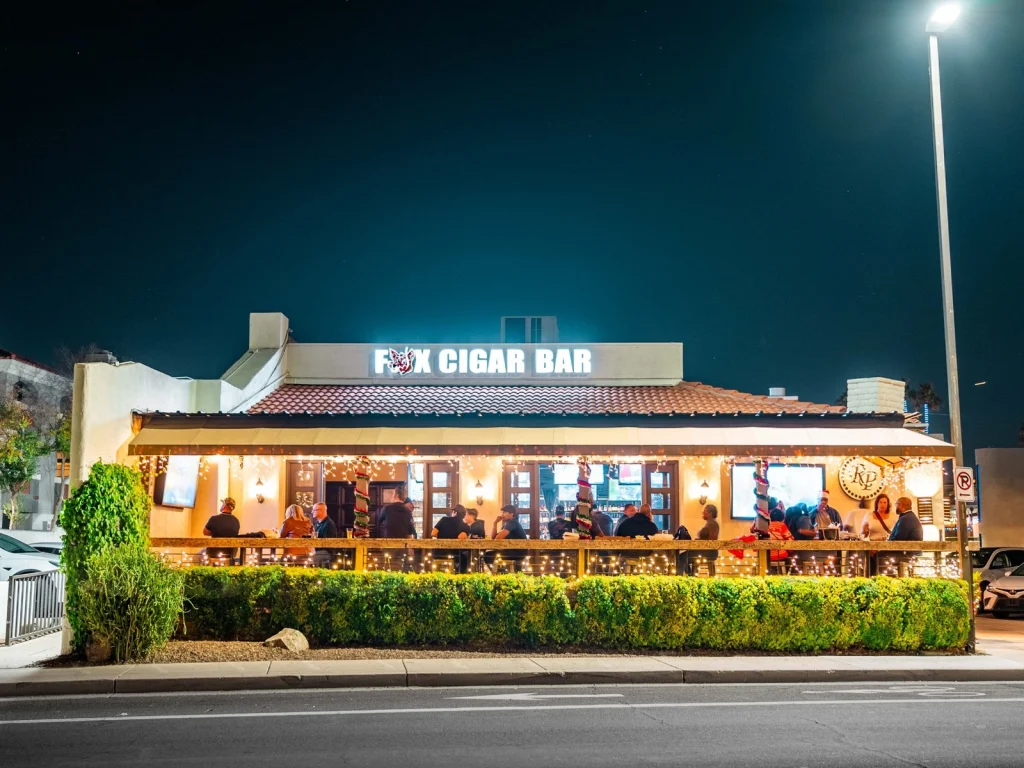 Fox Cigar Bar Scottsdale at night with its lit sign, patio full of guests and string lights behind a trimmed hedge