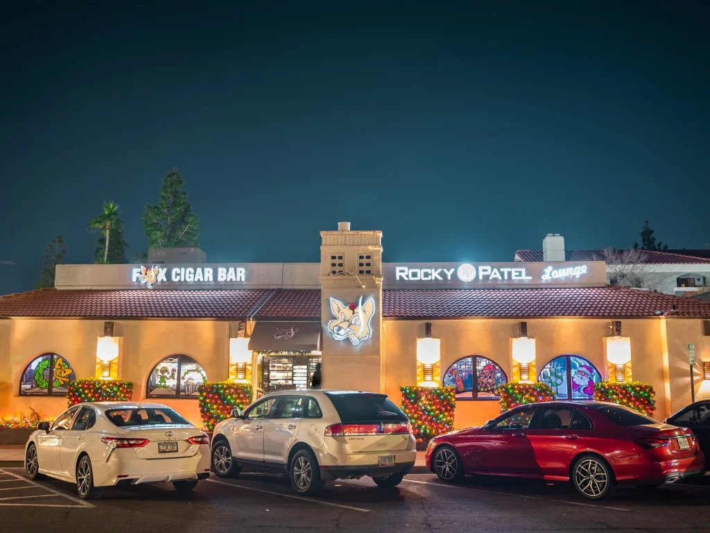 Storefront of Fox Cigar Bar Scottsdale and its Rocky Patel Lounge at night, holiday lights and painted windows, cars parked in front