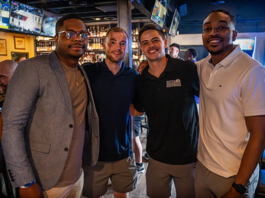 Four smiling friends posing together at Fox Cigar Bar Scottsdale, with TVs and a lit liquor shelf behind them