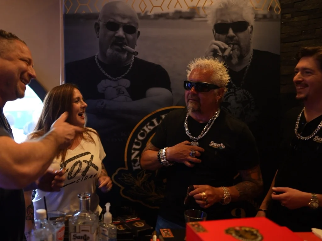 Guests laughing at a Santo cigar event at Fox Cigar Bar Scottsdale, in front of a Knuckle Sandwich banner