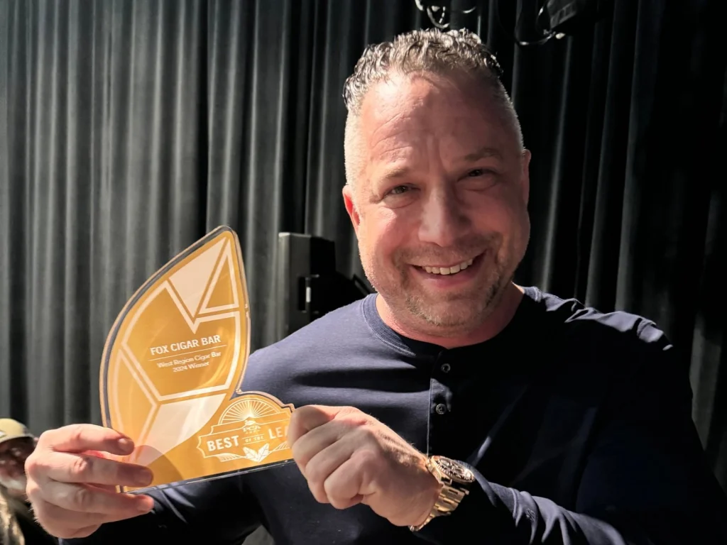 A smiling man holds the gold PCA Best of the Leaf award for Fox Cigar Bar, West Region Cigar Bar 2024 Winner